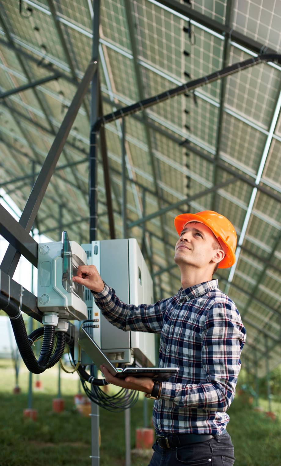 Operator in helmet and with laptop at station checking the operation of solar power plant. Vertical shot. Science technology. Solar panels. Global warming. Green energy.